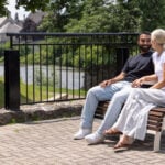 A man and a woman sit on a wooden ADA chair talking in a riverside park, part of the serene 720 Collection, with trees, grass, and a stone wall visible in the background.