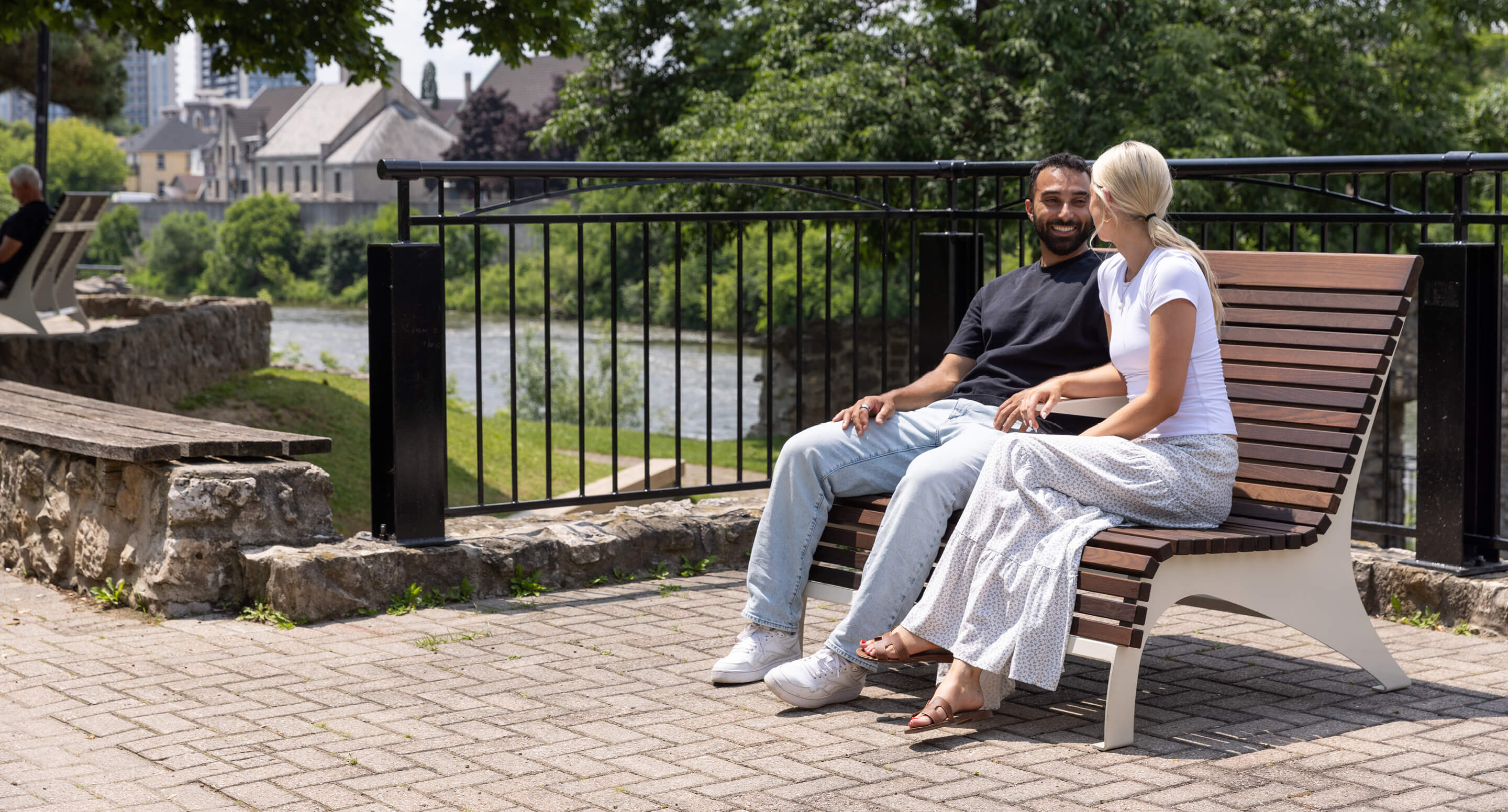 maglin-720-ADA-carousel A man and a woman sit on a wooden ADA chair talking in a riverside park, part of the serene 720 Collection, with trees, grass, and a stone wall visible in the background.
