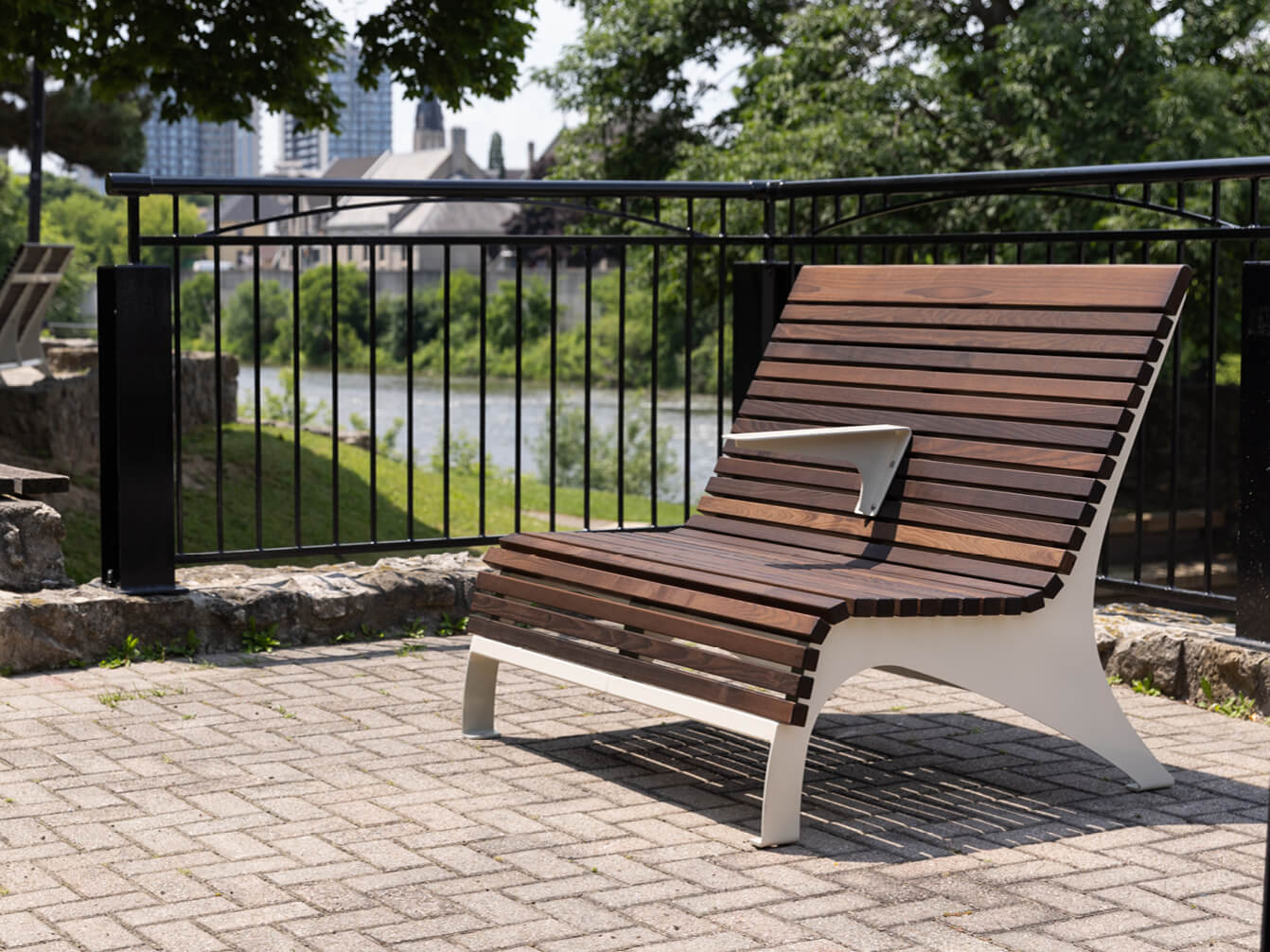 Maglin's 720 ADA Double Lounge Chair from the 720 Collection sits on a paved area near a black metal fence, with trees, water, and buildings in the background.