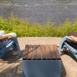 Two people sit in blue chairs from the 720 Collection, facing each other outside by a river, with a small table between them and greenery in the background.