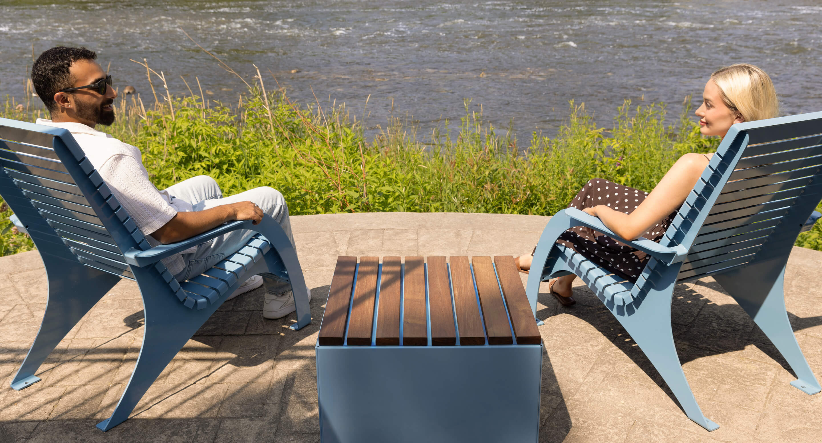 maglin-720-chairs-carousel Two people sit in blue chairs from the 720 Collection, facing each other outside by a river, with a small table between them and greenery in the background.