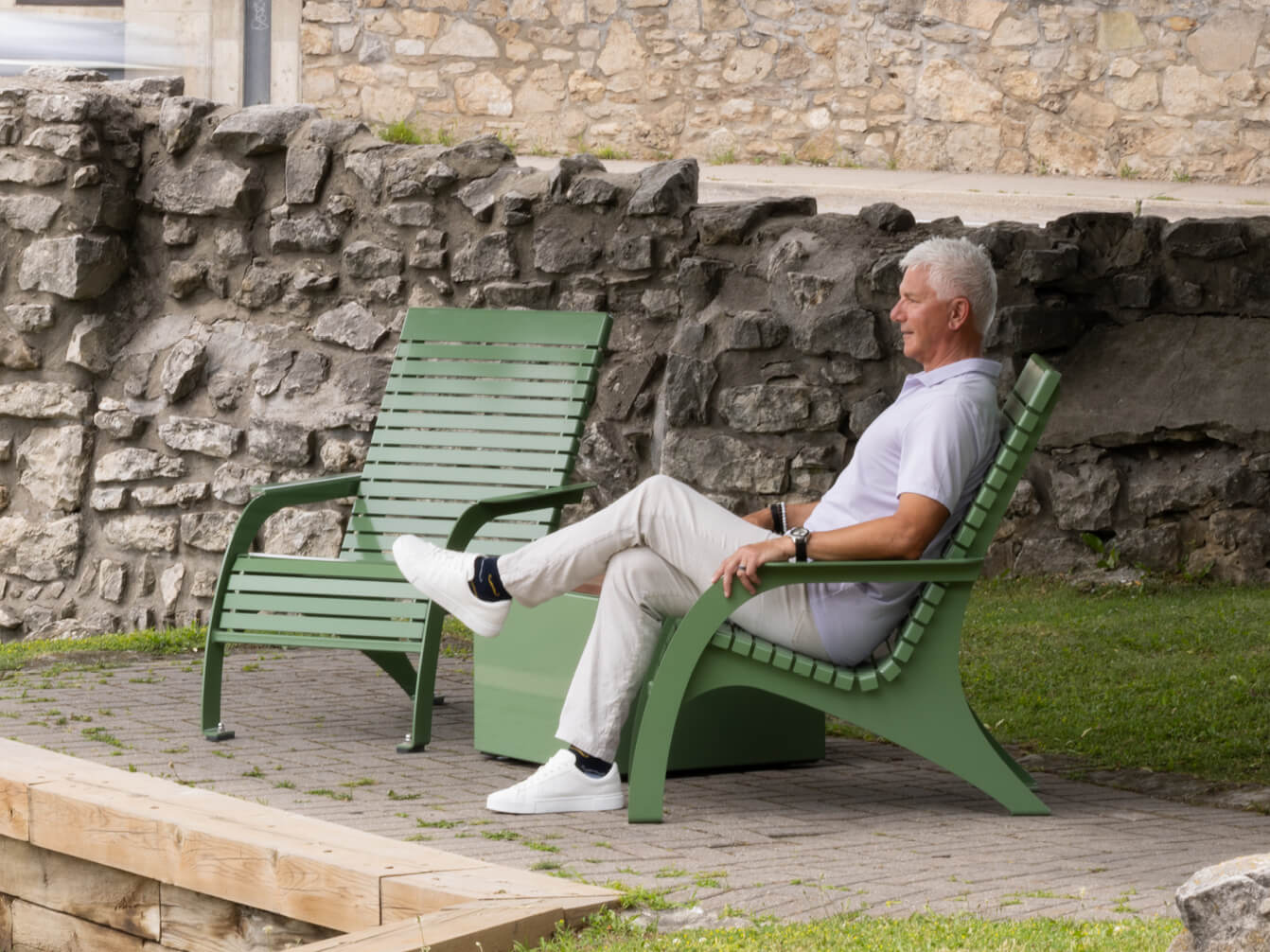 An older man in a white polo and light pants sits on a green outdoor bench near a stone wall, with his legs crossed and hands resting on his lap, embodying timeless style from the 720 Collection.