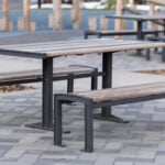 A wooden picnic table from the 720 Collection with two benches sits on a paved area outdoors, with another table in the background.