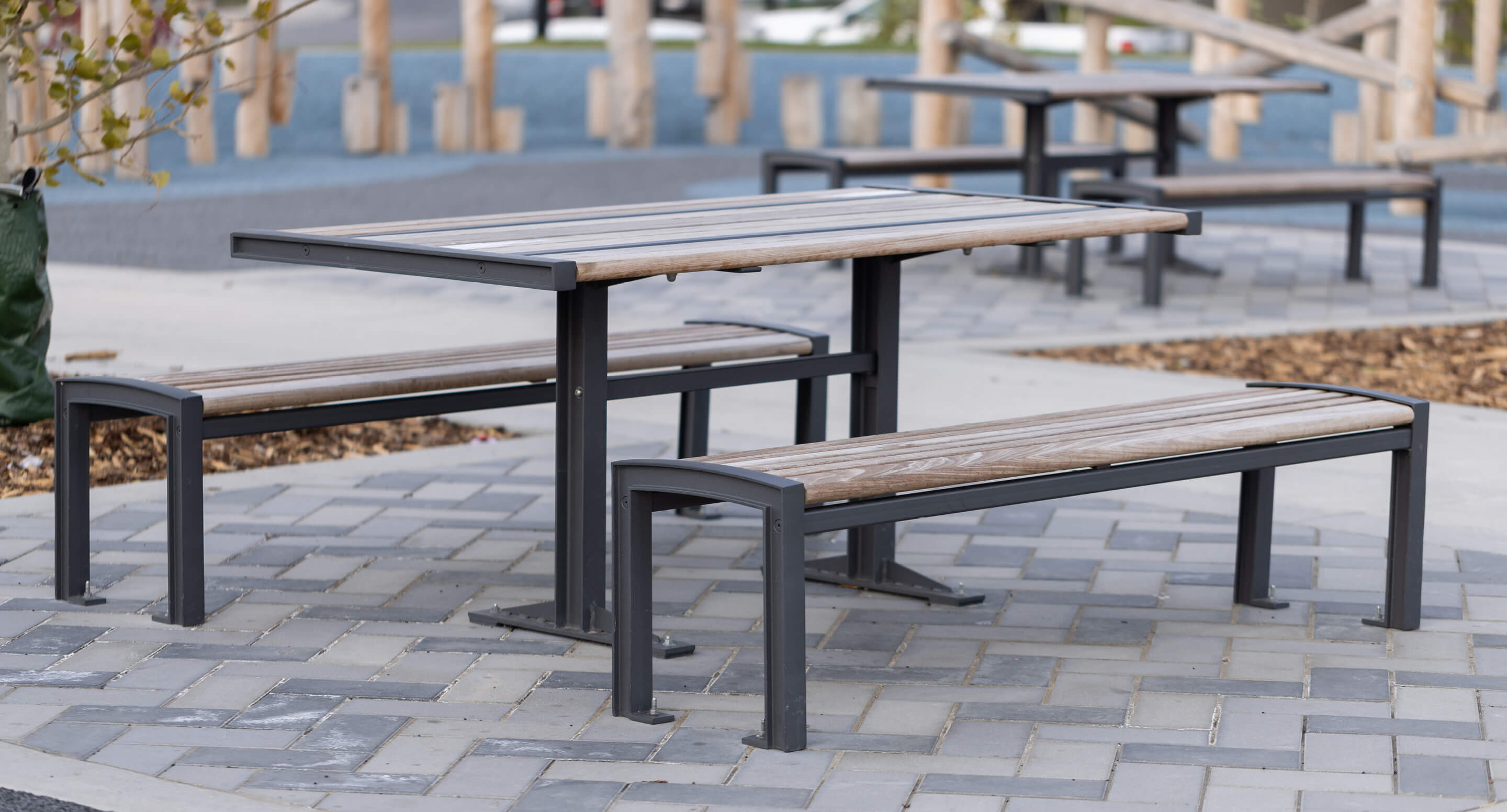 maglin-720-cluster-carousel A wooden picnic table from the 720 Collection with two benches sits on a paved area outdoors, with another table in the background.