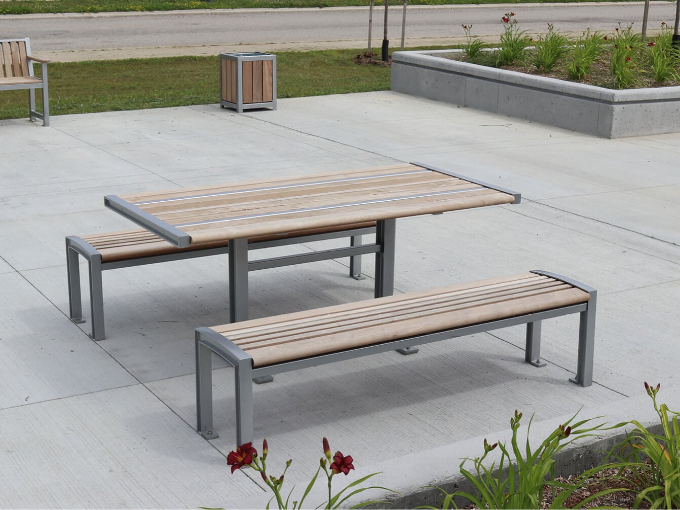 Outdoor wooden 720 picnic table from the 720 Collection with two matching benches on a concrete patio, surrounded by landscaping and a nearby road.