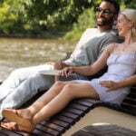 A man and a woman sit on a 720 double lounge chair by a river, smiling and enjoying the outdoors on a sunny day, creating an idyllic scene reminiscent of the 720 Collection.