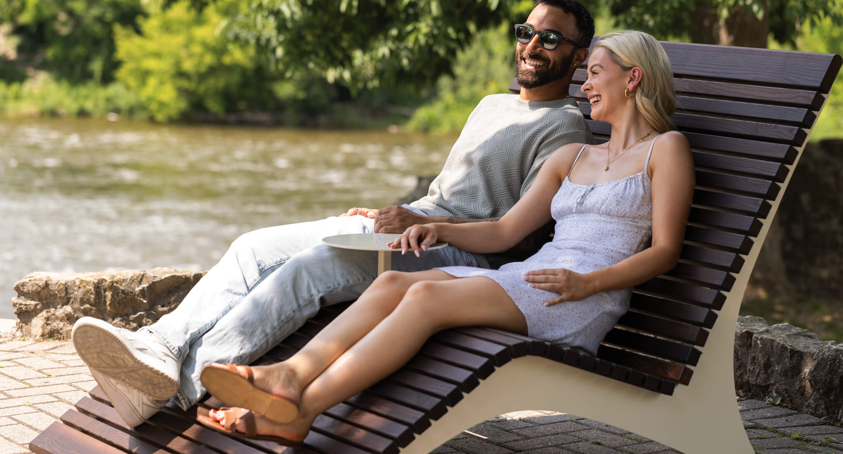 maglin-720-double-lounge-hero A man and a woman sit on a 720 double lounge chair by a river, smiling and enjoying the outdoors on a sunny day, creating an idyllic scene reminiscent of the 720 Collection.
