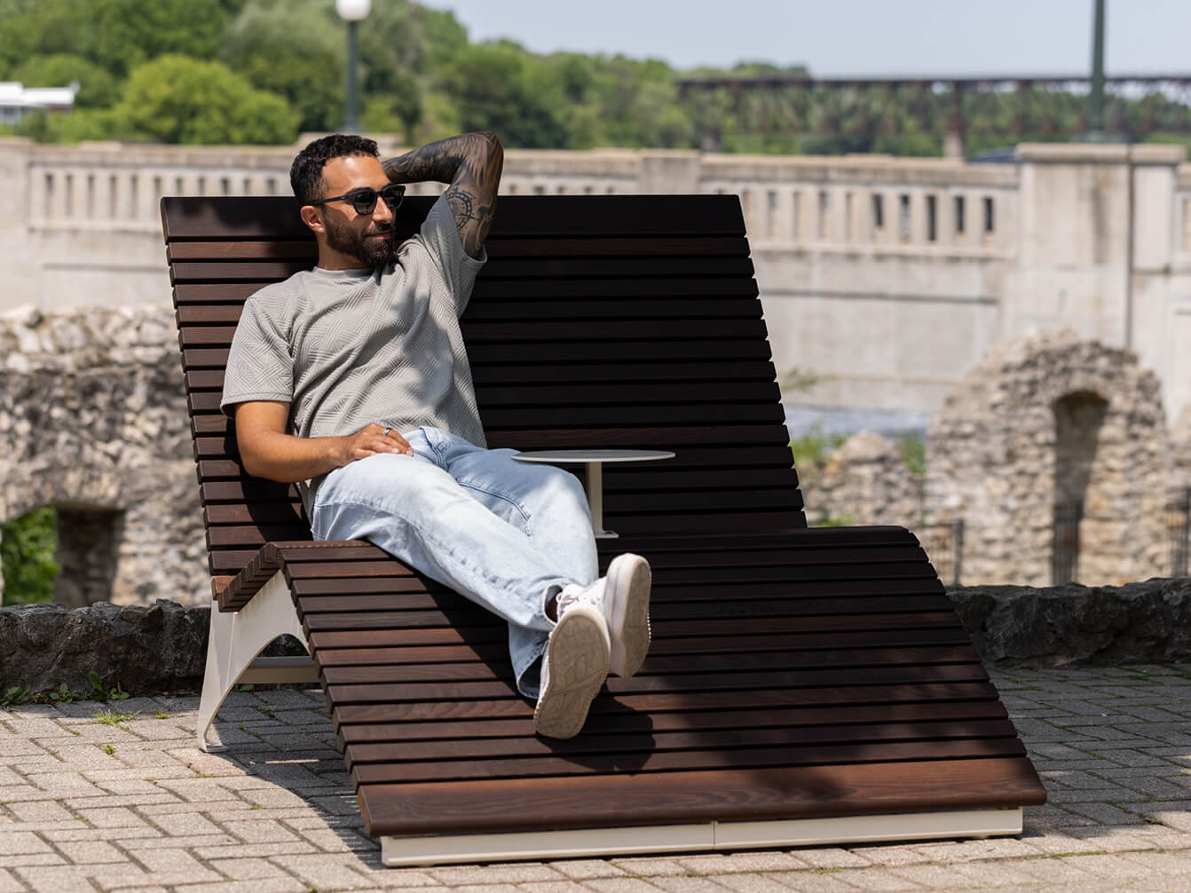 Man in sunglasses and casual clothing relaxes on a modern wooden outdoor double lounge chair from the 720 Collection beside stone ruins and greenery under a sunny sky.