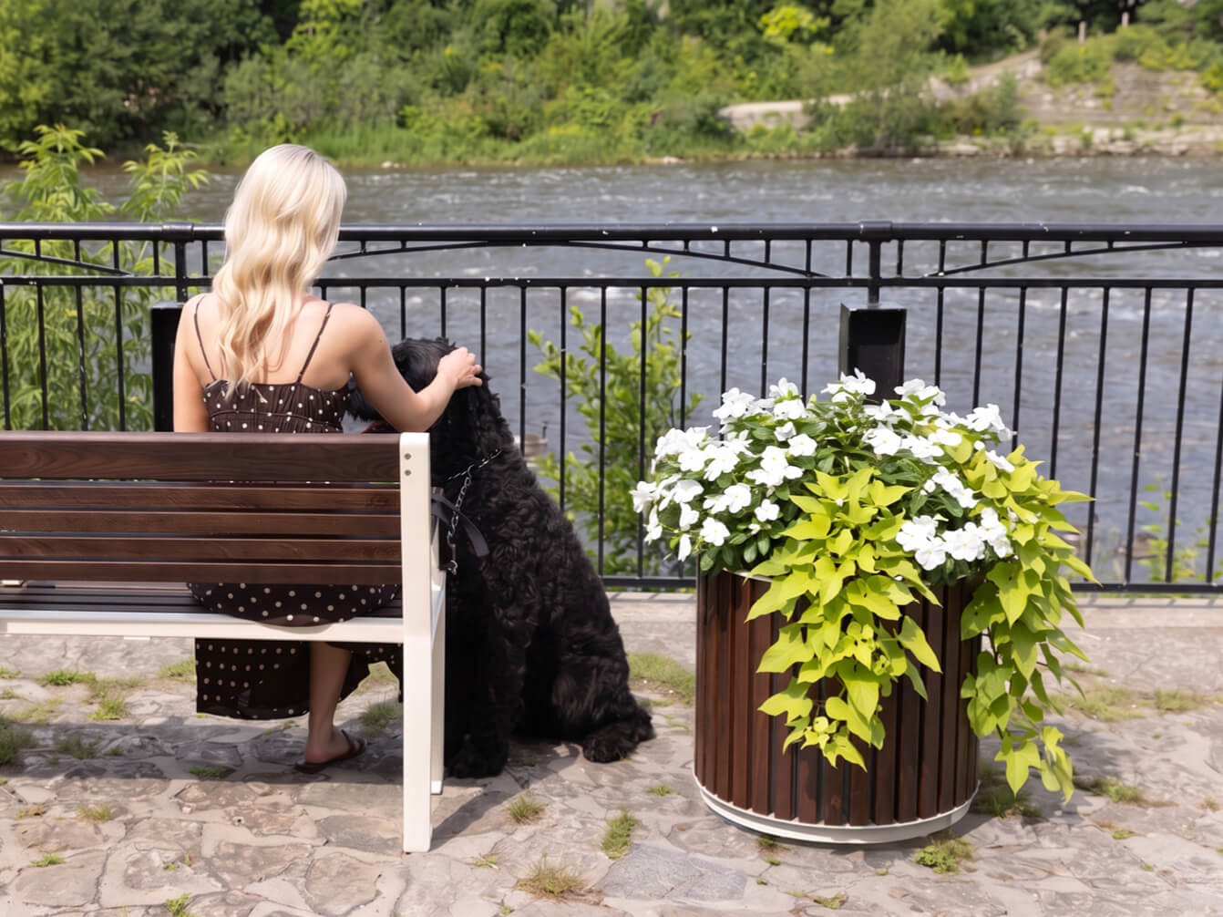 A woman sits on a bench by a river, her arm around a large black dog from the 720 Collection. A planter with white flowers and green leaves is nearby, while trees and a metal fence form the background.