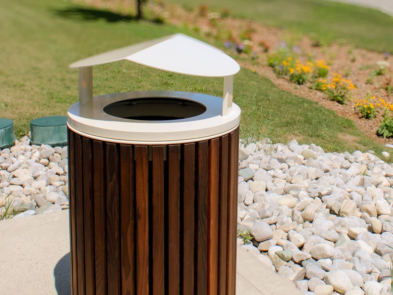 Outdoor trash can from the 720 Collection with a slanted white lid, situated on a concrete surface surrounded by rocks and grass with flowers in the background.