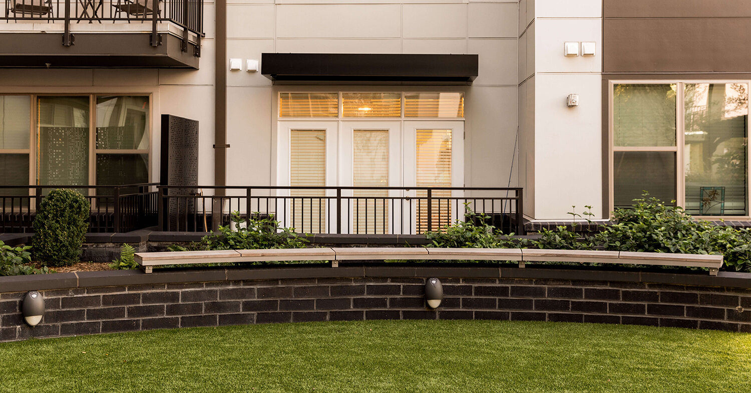 ogden-wall-mount-bench-link-mint-st-charlotte-nc-2 Curved wooden bench on artificial grass in front of a modern apartment building with white patio doors and railings at Link Apartments on Mint Street in Charlotte, NC.