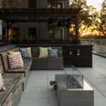 A modern outdoor patio at Link Apartments on Mint Street in Charlotte, NC, featuring wooden benches, colorful cushions, a fire pit, and a wooden pergola at sunset.