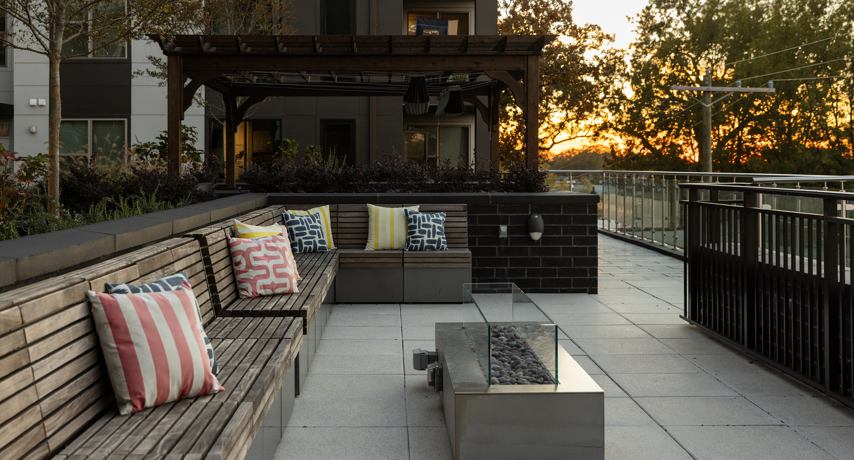 A modern outdoor patio at Link Apartments on Mint Street in Charlotte, NC, featuring wooden benches, colorful cushions, a fire pit, and a wooden pergola at sunset.