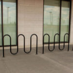 A black metal bike rack with a wave design is installed on a concrete sidewalk in front of large windows with blinds at Groves Memorial Community Hospital.
