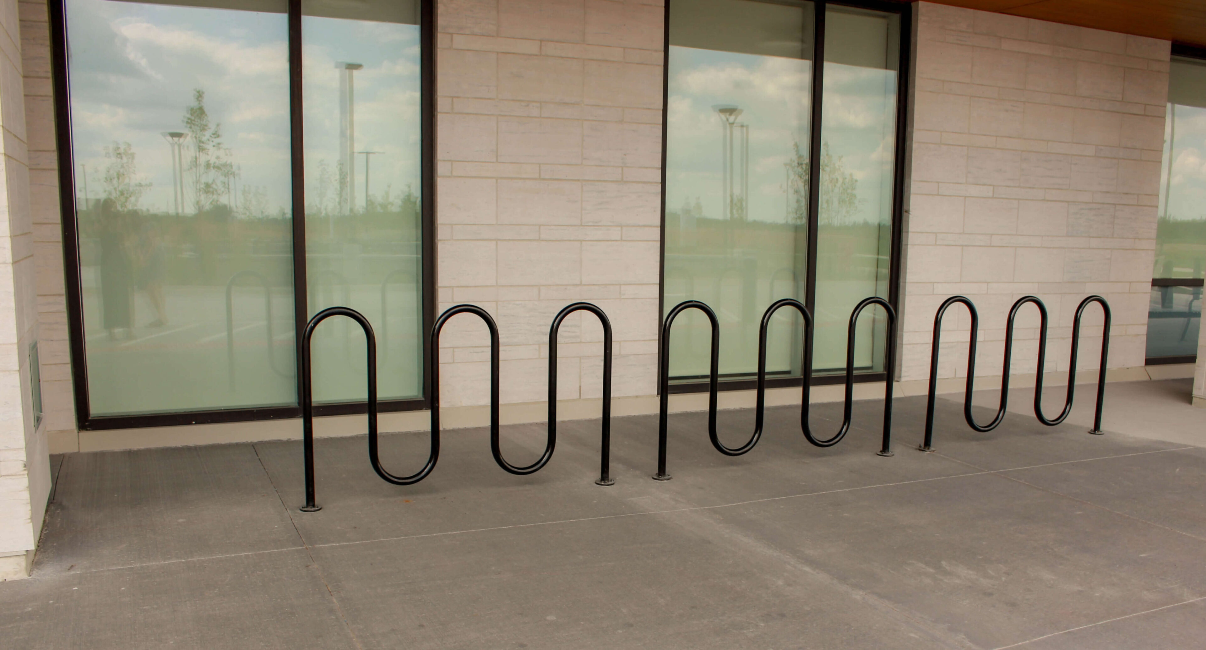 groves-memorial-hospital-400-wave-bike-rack A black metal bike rack with a wave design is installed on a concrete sidewalk in front of large windows with blinds at Groves Memorial Community Hospital.