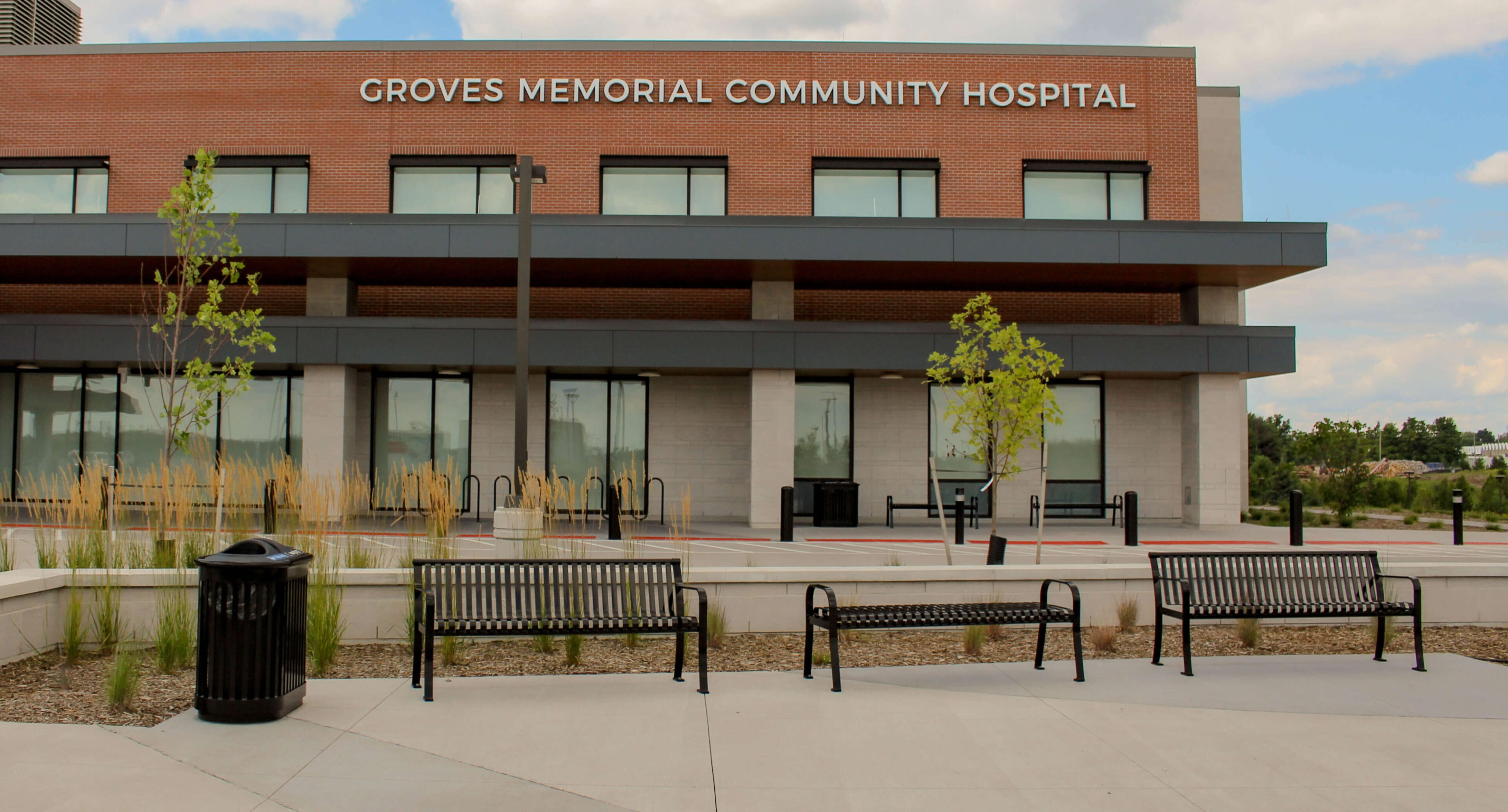 groves-memorial-hospital-510-backed-backless-bench-250-recycling Exterior view of Groves Memorial Community Hospital showcasing the hospital’s brick building, benches, trees, and bike racks on a clear day. Groves Memorial Community Hospital’s welcoming exterior creates a peaceful and inviting atmosphere for patients and visitors alike.