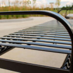 Close-up view of a black metal slatted bench in an outdoor park setting near Groves Memorial Community Hospital, with blurred greenery and sidewalk in the background.