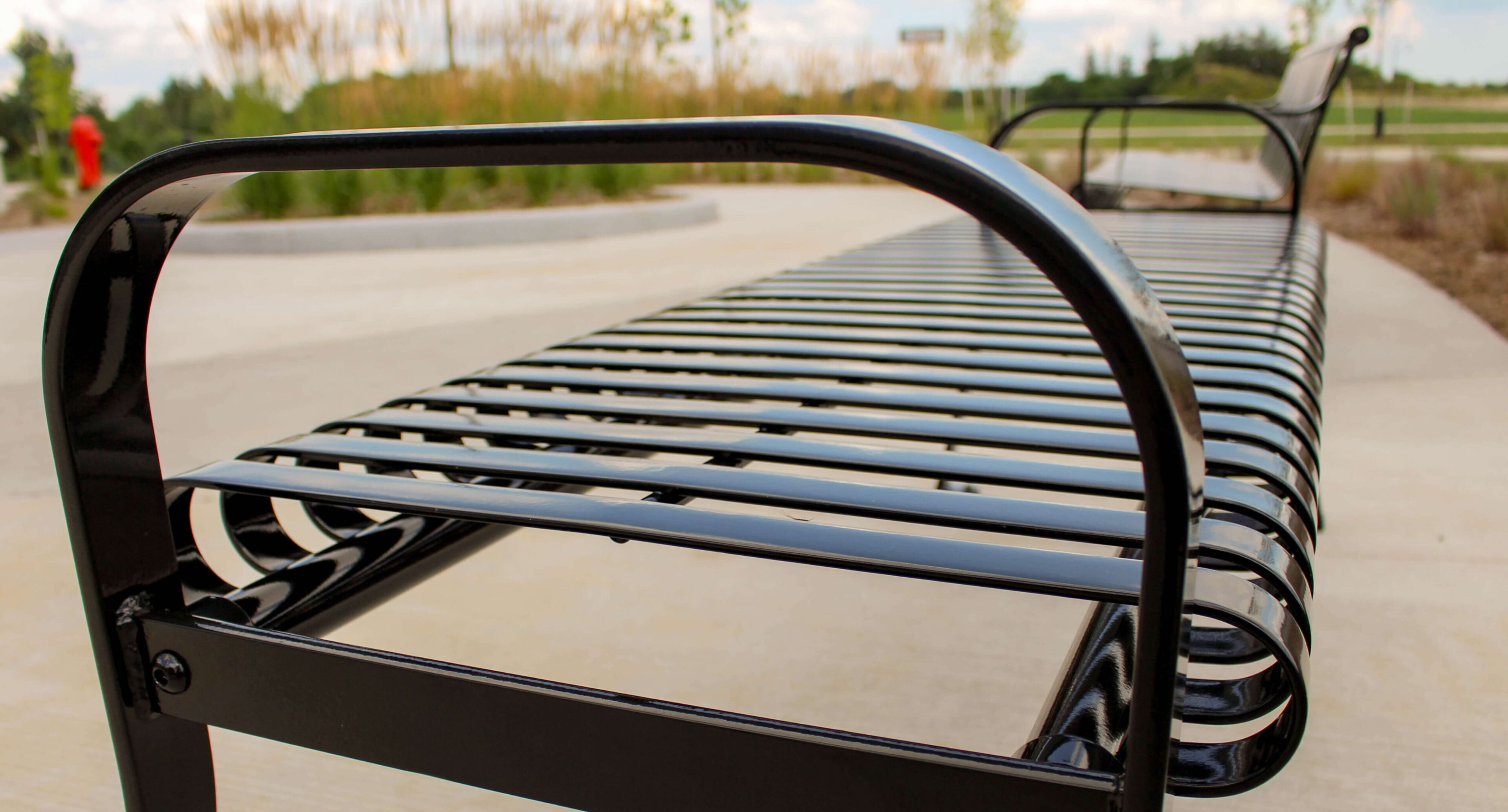 groves-memorial-hospital-510-backed-backless-bench-close Close-up view of a black metal slatted bench in an outdoor park setting near Groves Memorial Community Hospital, with blurred greenery and sidewalk in the background.