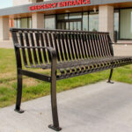 A black metal bench is placed on a concrete slab in front of Groves Memorial Community Hospital’s emergency entrance, with grass and a parking lot visible nearby.