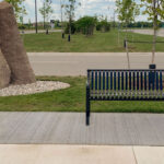 A black metal bench sits on a concrete pad beside a large stone and young trees near the parking lot of Groves Memorial Community Hospital on a cloudy day.