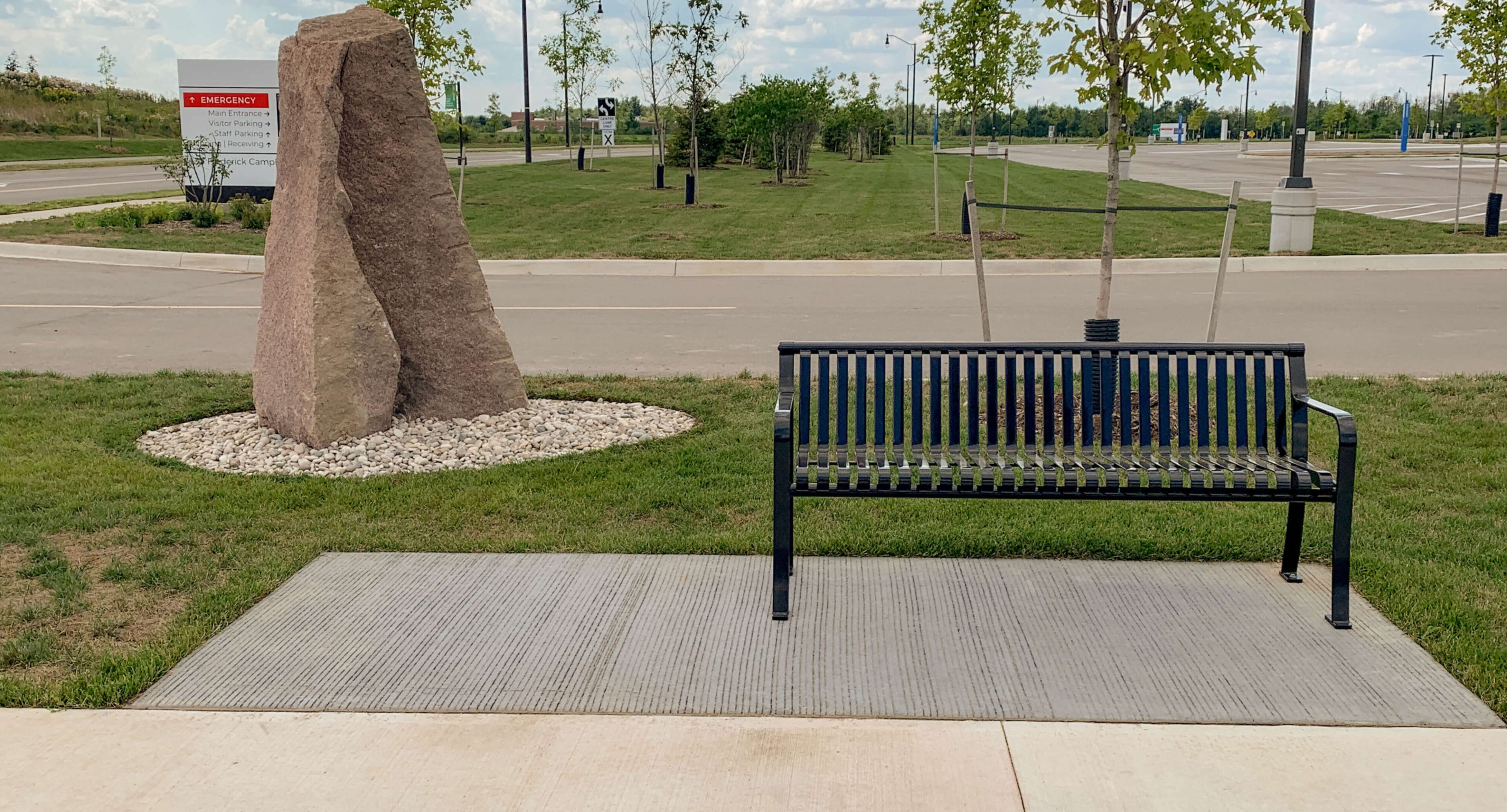 groves-memorial-hospital-510-backed-bench-2 A black metal bench sits on a concrete pad beside a large stone and young trees near the parking lot of Groves Memorial Community Hospital on a cloudy day.