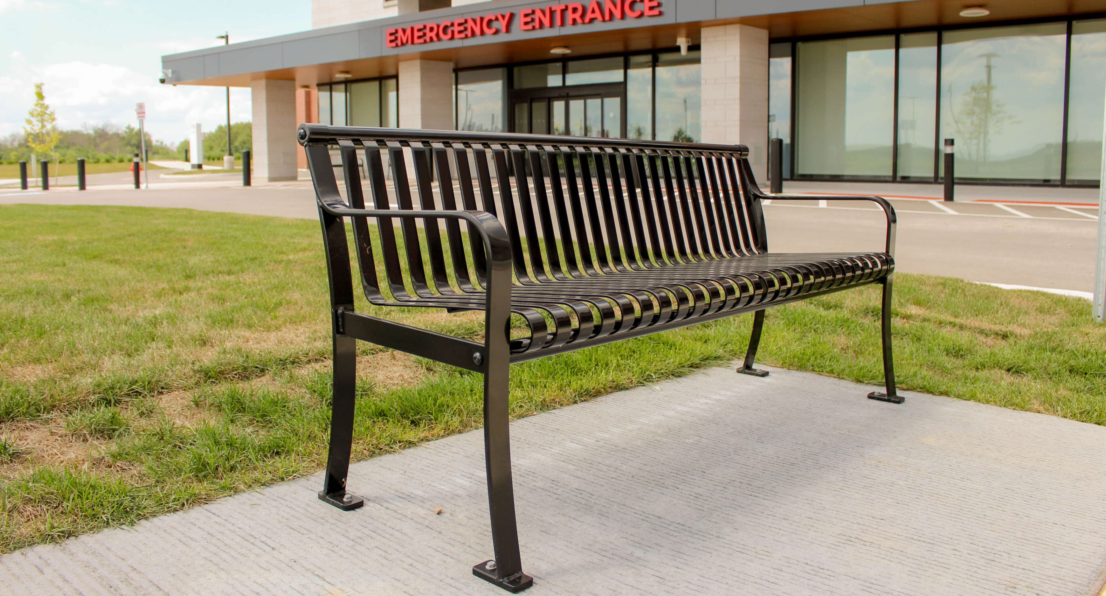 groves-memorial-hospital-510-backed-bench A black metal bench is placed on a concrete slab in front of Groves Memorial Community Hospital’s emergency entrance, with grass and a parking lot visible nearby.