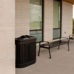 Two black metal benches and a trash can are placed outside Groves Memorial Community Hospital, set against the building’s light stone walls and large windows on a concrete patio.