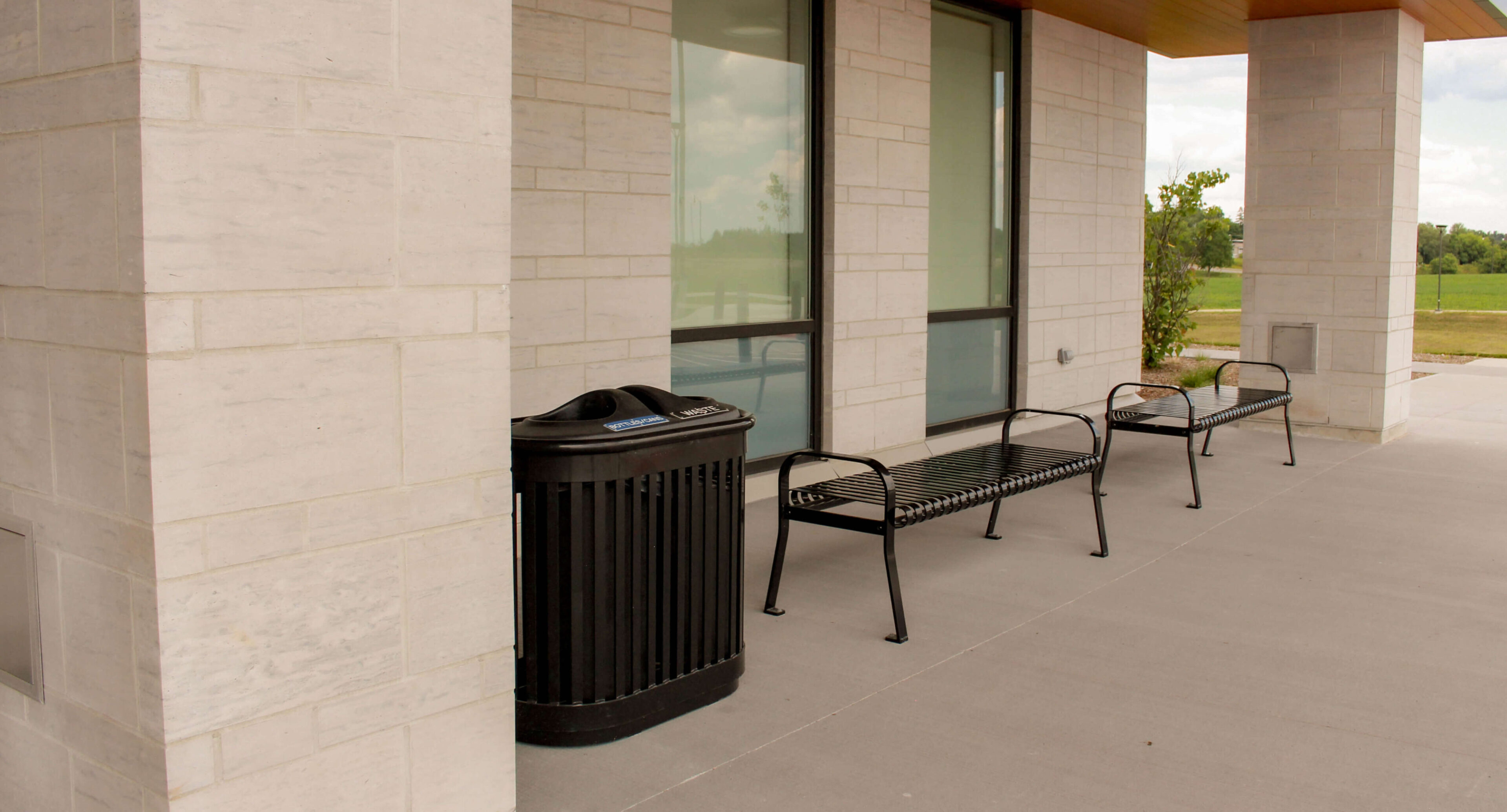 groves-memorial-hospital-entrance-250-recycling-510-backless-bench Two black metal benches and a trash can are placed outside Groves Memorial Community Hospital, set against the building’s light stone walls and large windows on a concrete patio.