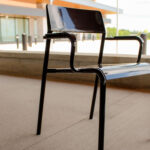 Black metal chair with armrests and a slightly angled backrest placed on a concrete patio outside Groves Memorial Community Hospital’s modern building.