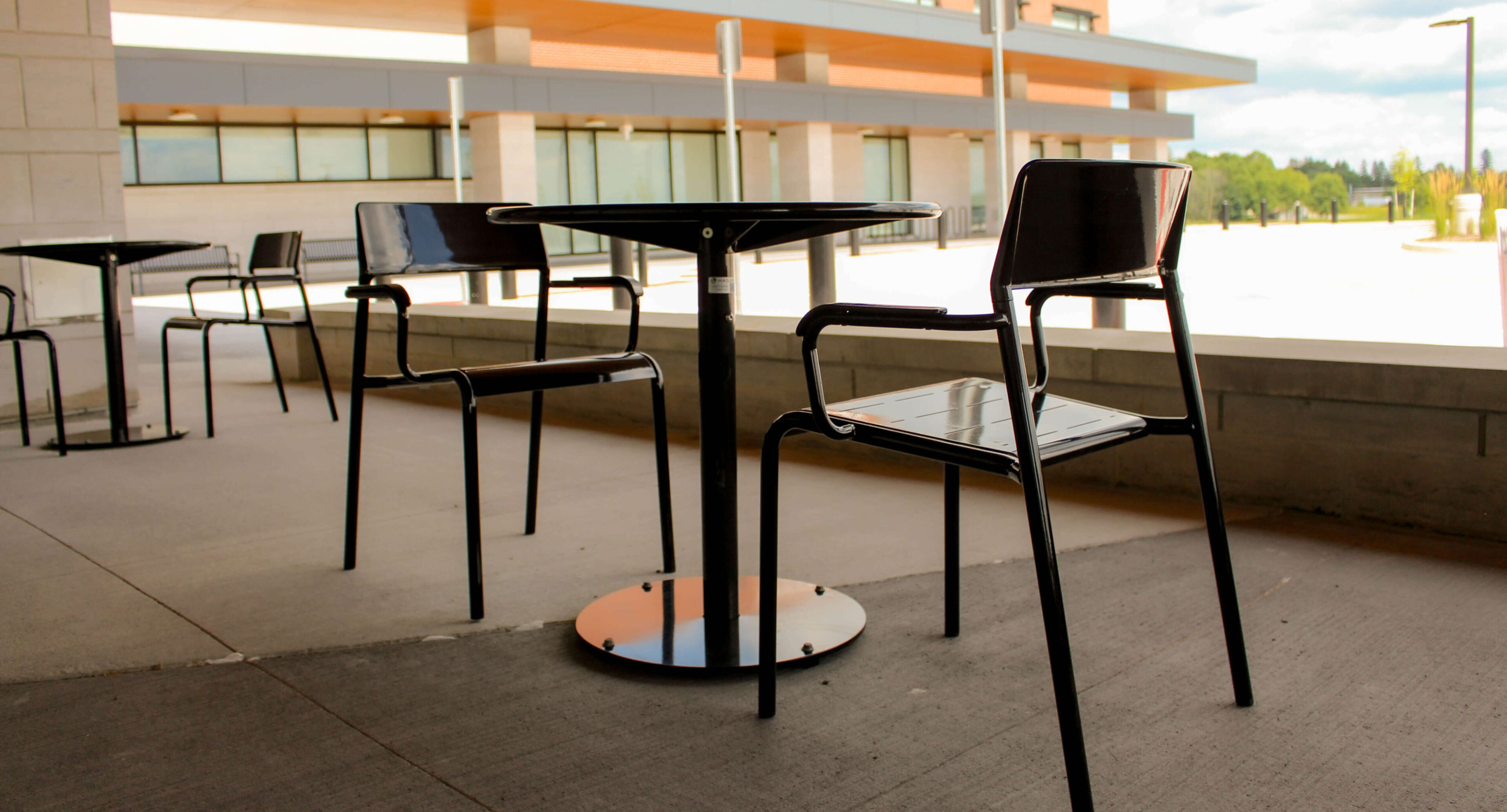groves-memorial-hospital-foro-tables-chairs Two black metal chairs and a round table are placed on a concrete outdoor patio at Groves Memorial Community Hospital, with a modern building and windows visible in the background.