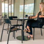 A woman in black sits alone at an outdoor café table with a drink in hand, just steps away from Groves Memorial Community Hospital. Empty tables and chairs surround her, with glass windows and a patio setting visible in the background.