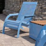 Light blue metal patio chair and matching storage box with a wooden top are on a stone-paved patio next to a stone wall.