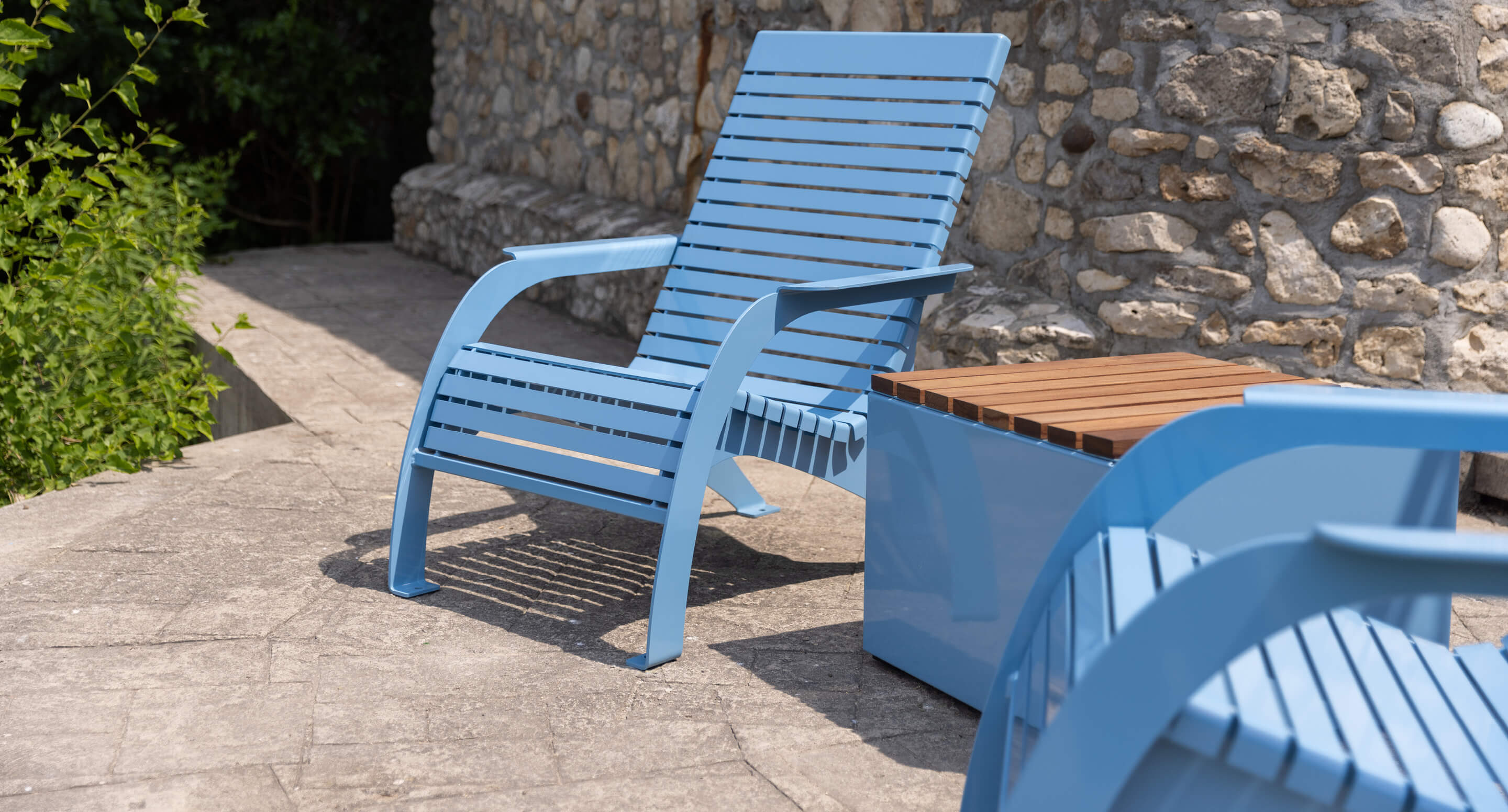 maglin-mch-0720-lounge-chair-blue Light blue metal patio chair and matching storage box with a wooden top are on a stone-paved patio next to a stone wall.