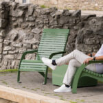 An older man in light-colored clothes sits on a green outdoor chair near a stone wall, with an empty chair beside him.