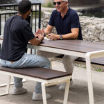 Two men sit at a wooden outdoor table near a river, talking. One wears a black shirt and jeans, the other a navy shirt and light pants. A metal fence and stone wall are in the background.