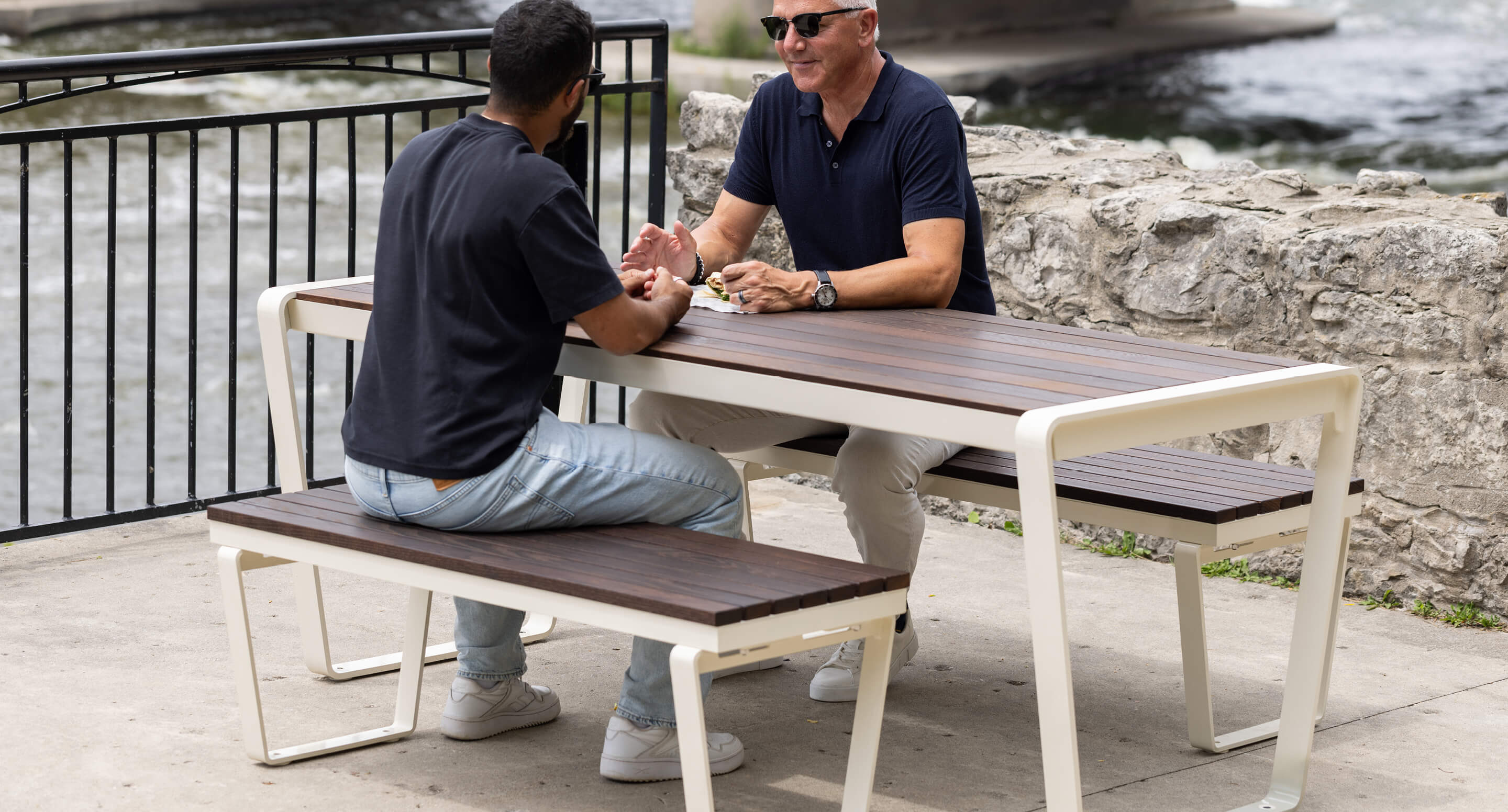 Two men sit at a wooden outdoor table near a river, talking. One wears a black shirt and jeans, the other a navy shirt and light pants. A metal fence and stone wall are in the background.
