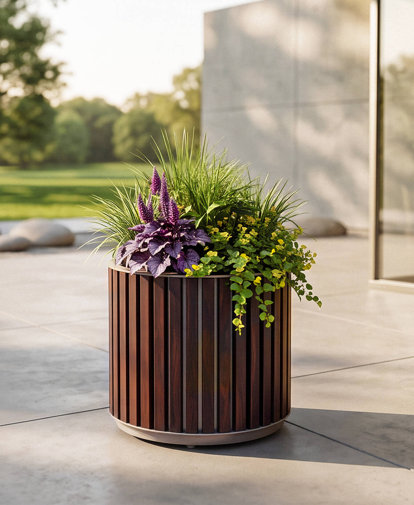 Round wooden planter with green and purple plants sits on a patio near a modern building; lawn and trees are visible in the background.