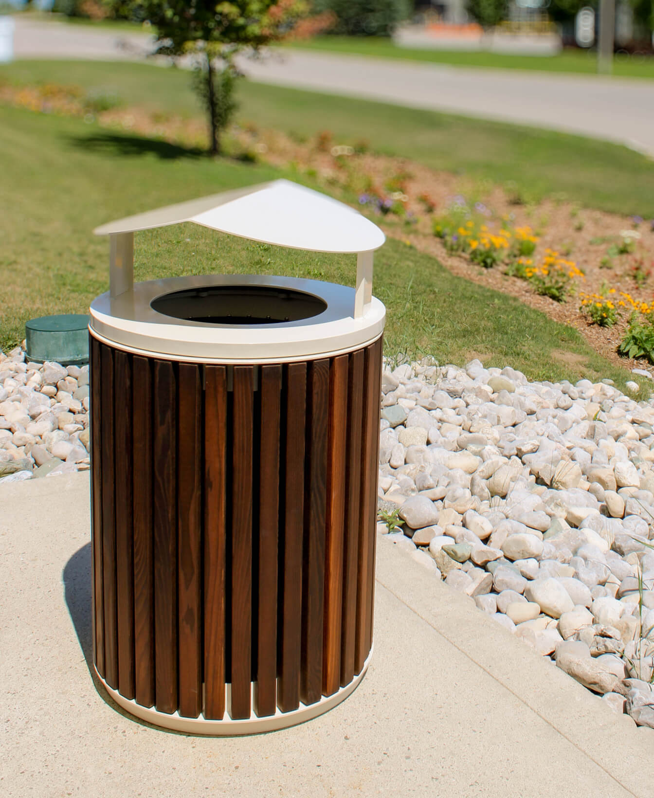 Outdoor trash can with vertical wooden slats and a white, angled metal lid, placed on a sidewalk next to rocks and landscaping with flowers and grass.