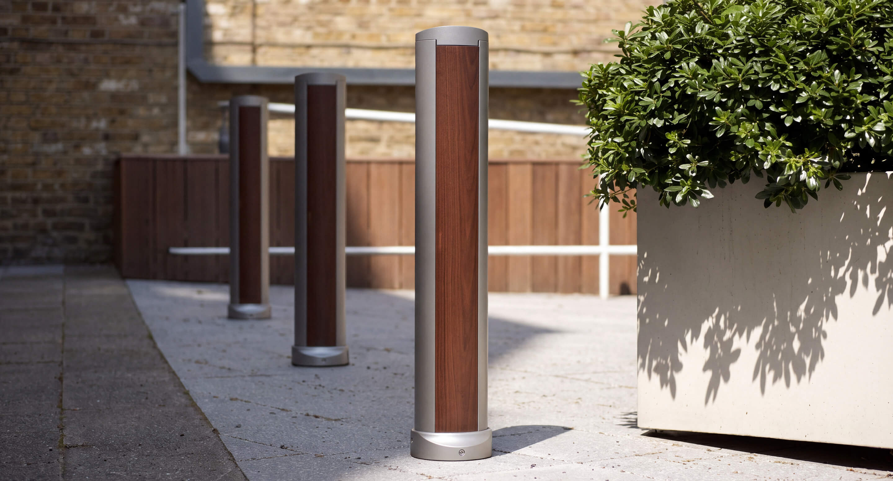 maglin_mbo-0720-bollards_carousel Trois bornes cylindriques ornées de panneaux en bois sont disposées le long d'une allée pavée en plein air, à côté d'un grand bac à fleurs contenant un arbuste au feuillage vert. Un mur en briques et une balustrade se dessinent en arrière-plan.
