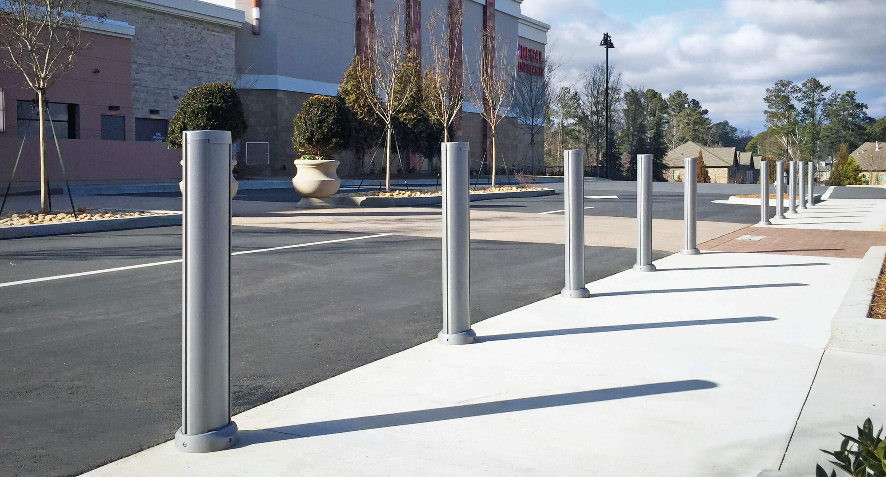maglin_mbo-0720-bollards_carousel2 Une rangée de bornes métalliques borde le trottoir près d'un grand immeuble commercial, avec en arrière-plan un parking presque vide.