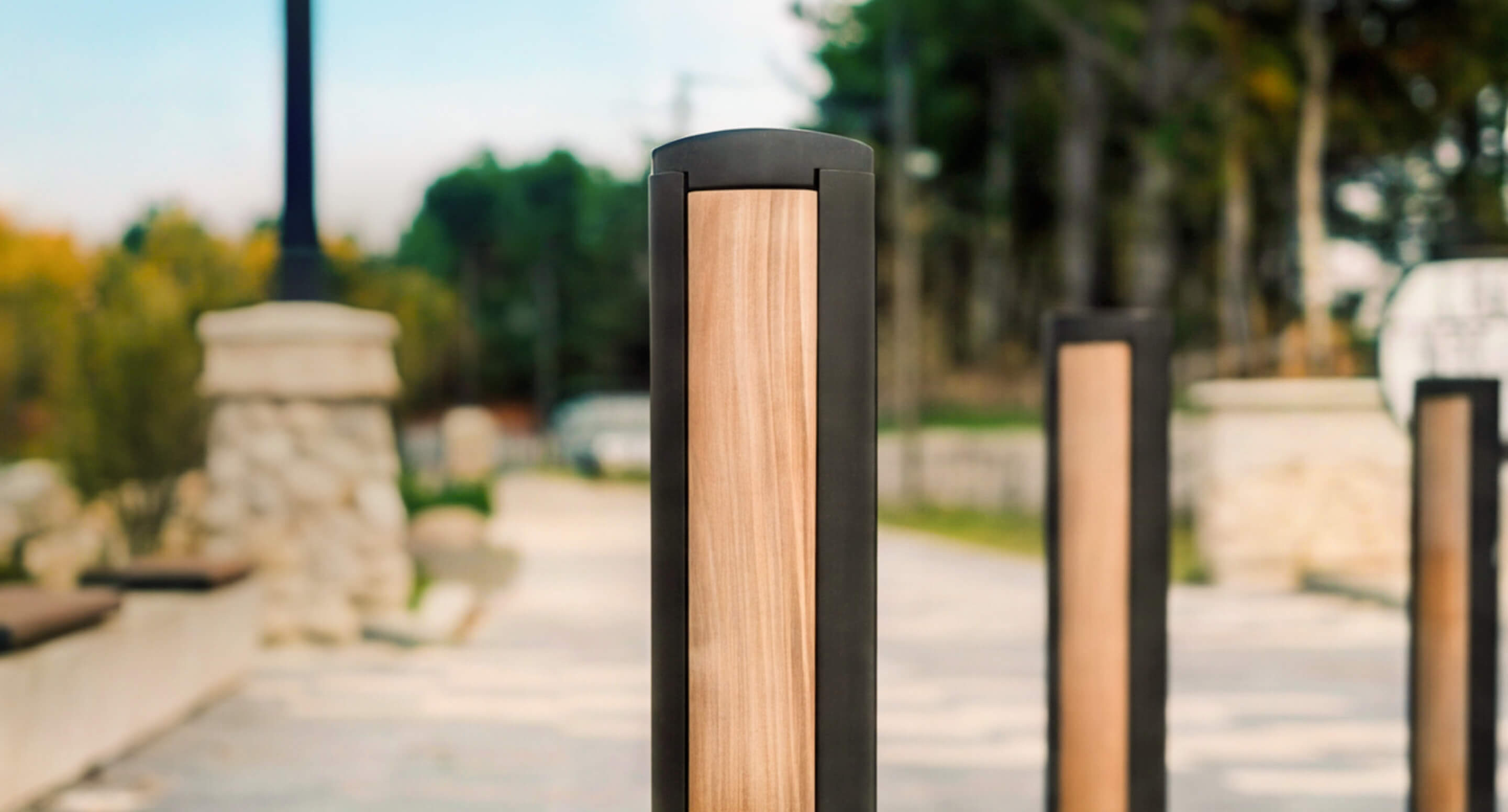 maglin_mbo-0720-bollards_carousel3 Gros plan sur une borne moderne en bois et en métal, située sur une allée extérieure pavée, bordée d'arbres, avec des structures en pierre en arrière-plan.