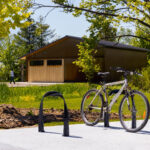 A bicycle is locked to a bike rack on a concrete pad in a park, with a wooden building, trees, grass, and a walking path in the background.