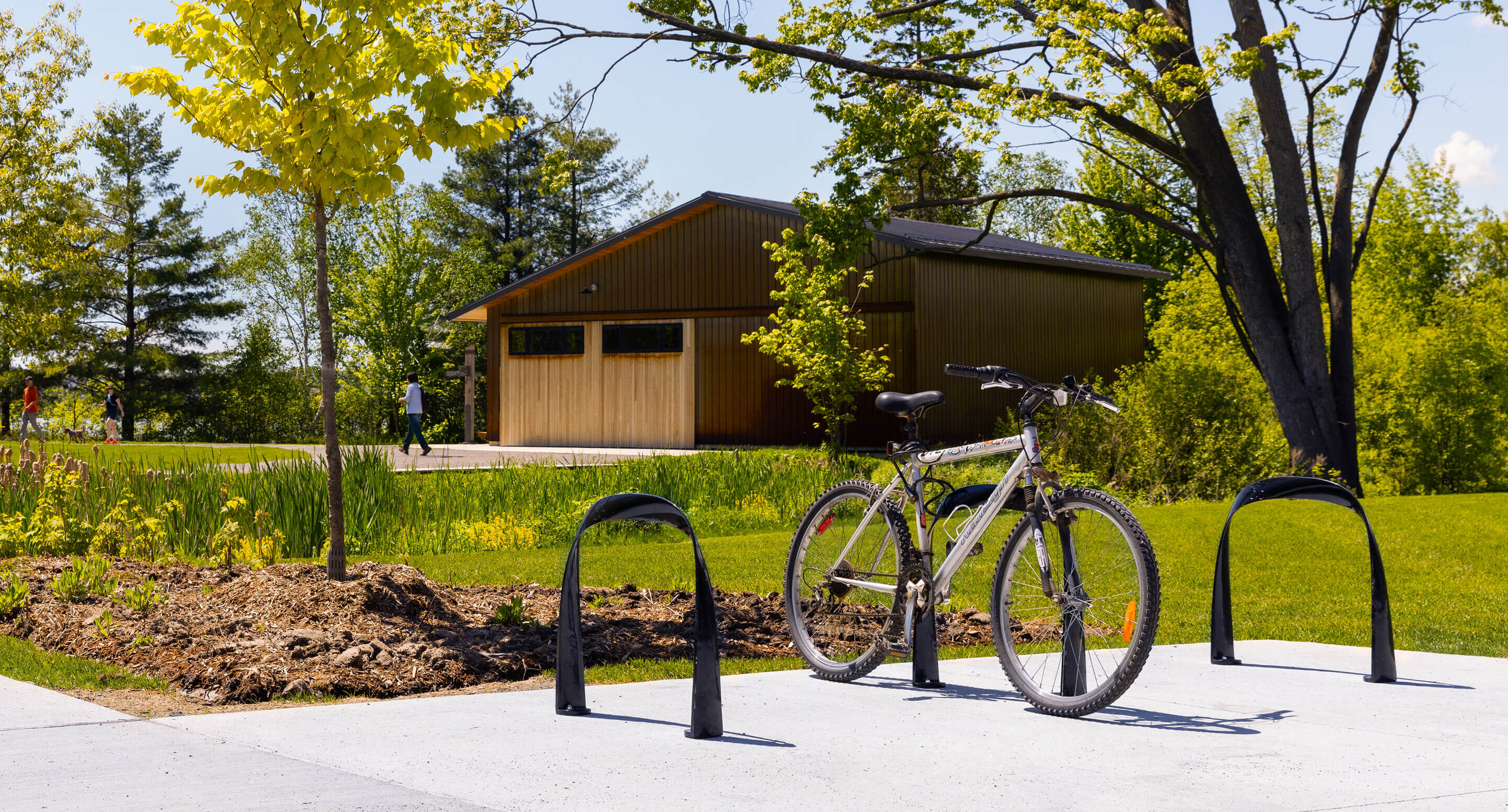 maglin_mbr-0720-bike-racks_carousel A bicycle is locked to a bike rack on a concrete pad in a park, with a wooden building, trees, grass, and a walking path in the background.