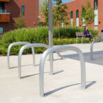 Empty metal bike racks on a concrete path outside a modern brick building; a person sits on a bench in the background near landscaped greenery.