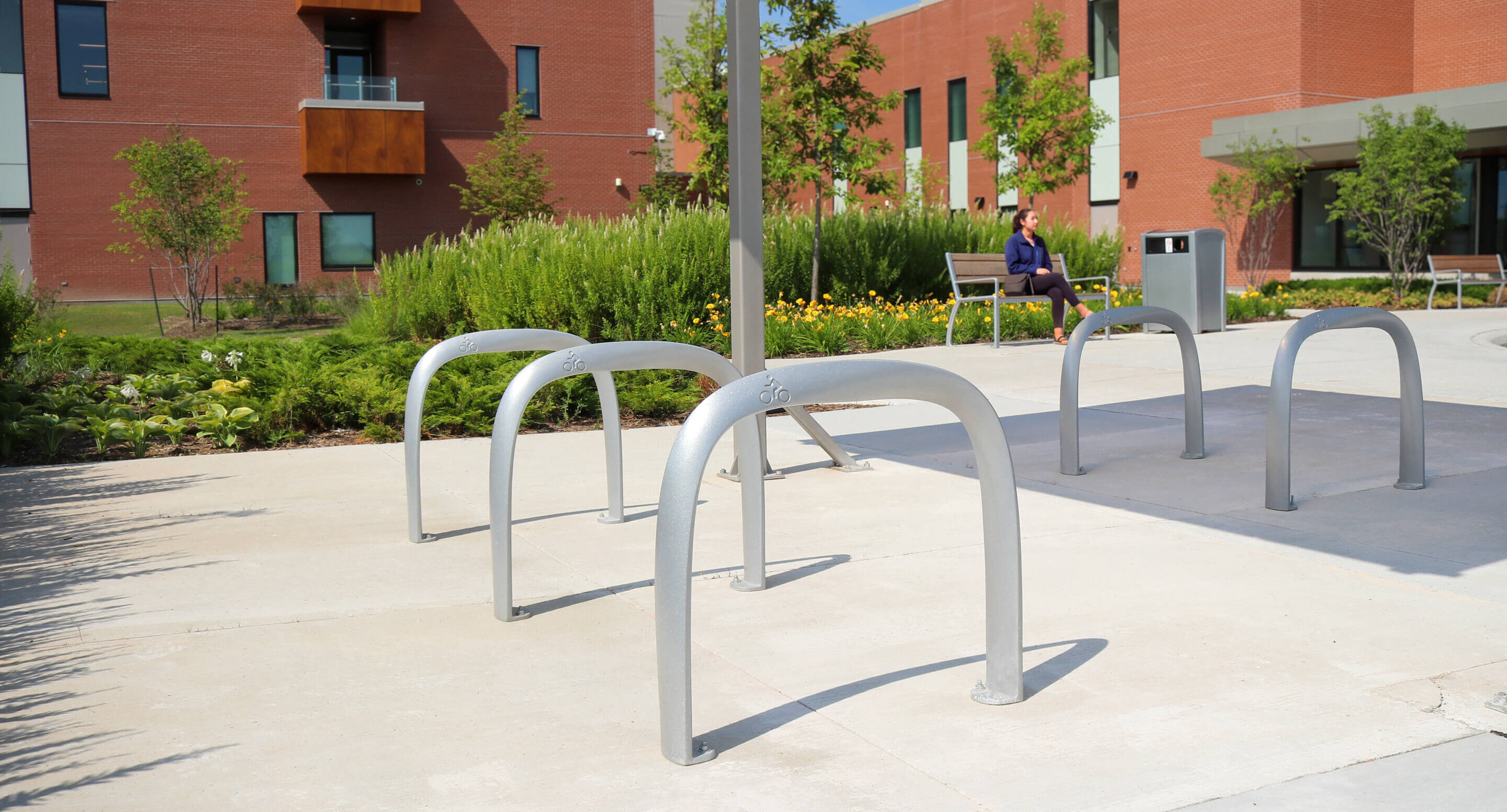 maglin_mbr-0720-bike-racks_carousel2 Empty metal bike racks on a concrete path outside a modern brick building; a person sits on a bench in the background near landscaped greenery.
