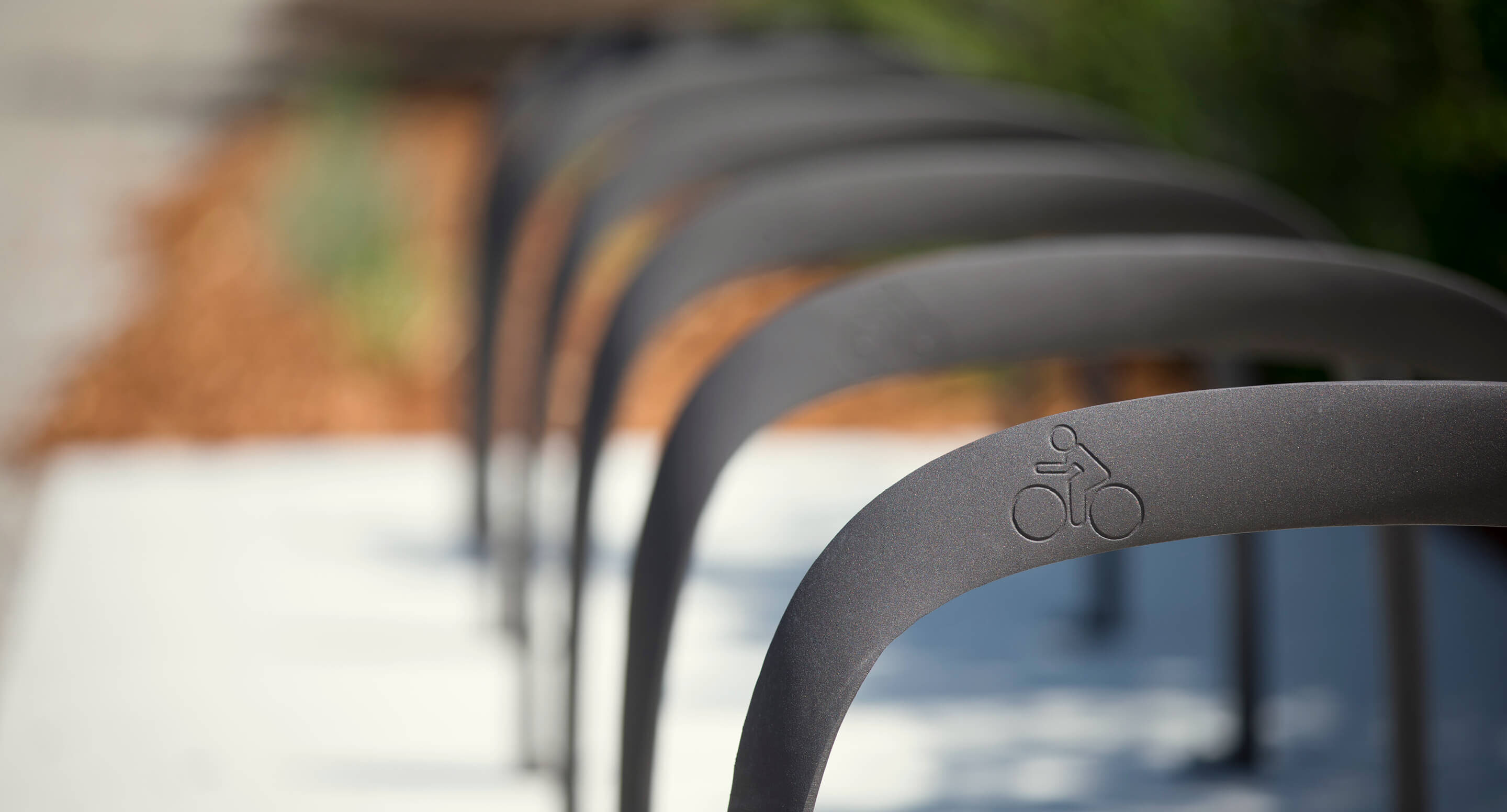 maglin_mbr-0720-bike-racks_carousel3 Close-up of a row of metal bicycle racks with a bike symbol engraved on the nearest one, positioned outdoors on a paved surface.