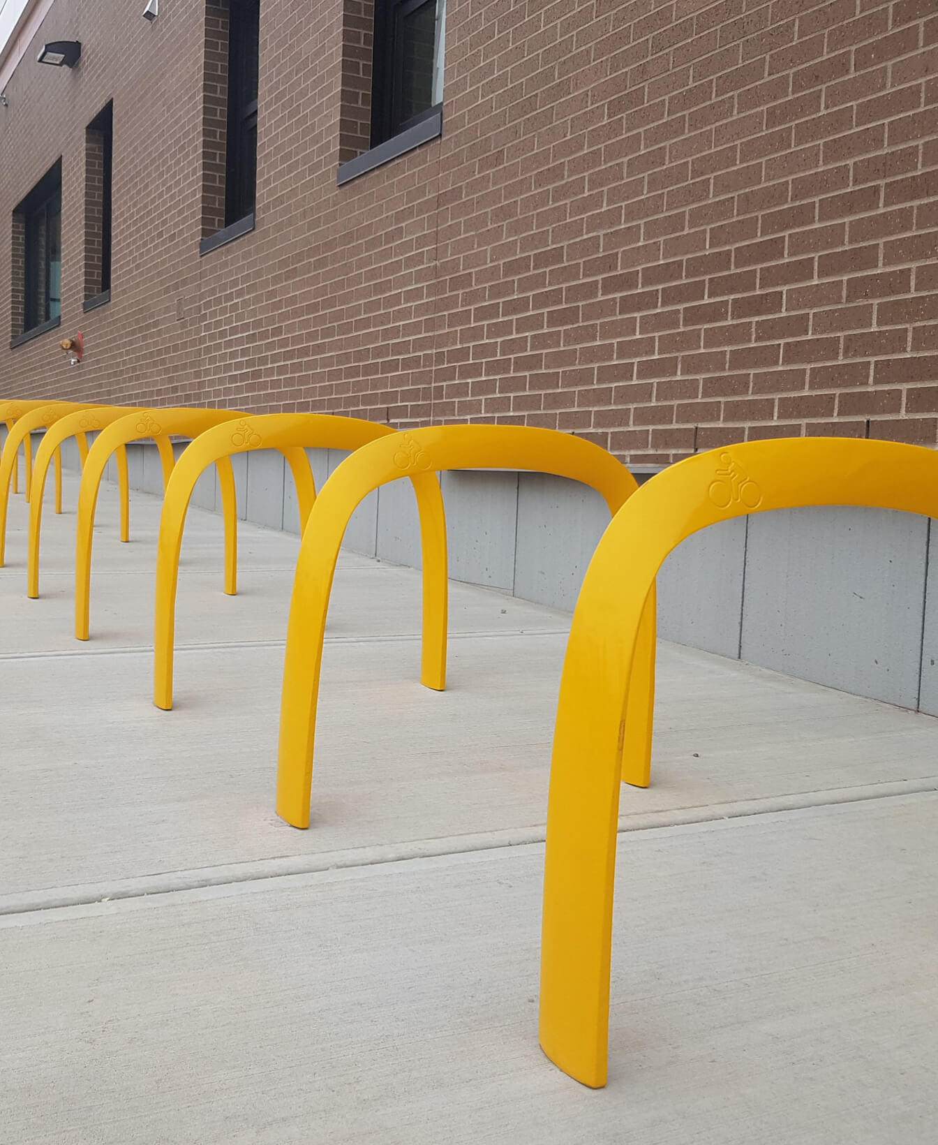 A row of yellow bike racks is installed on a concrete sidewalk next to a brick building with several windows.
