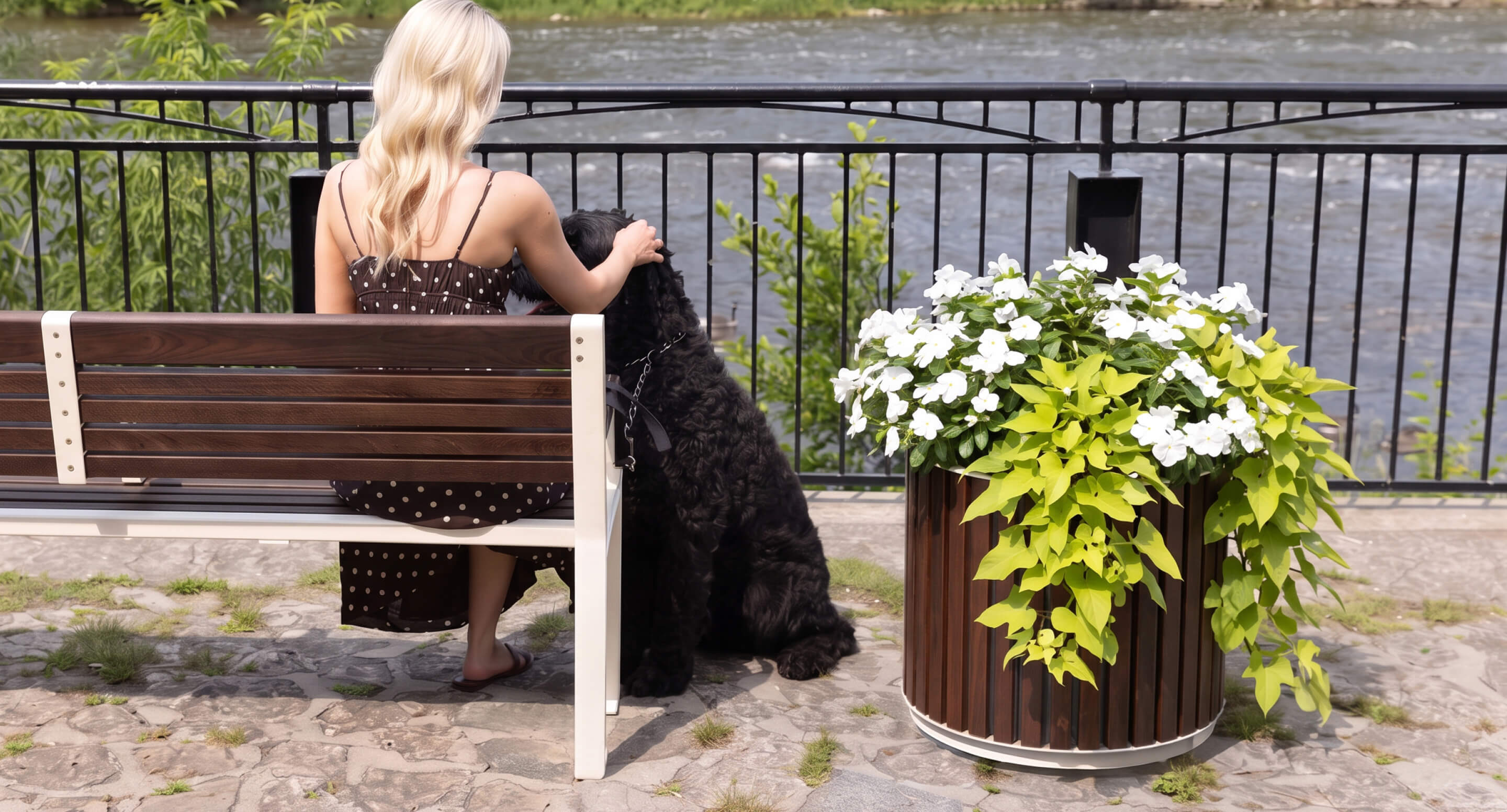 maglin_mpl-0720-planters_carousel A woman with blonde hair sits on a bench by a river, petting a large black dog. A planter with white flowers and green leaves is nearby.