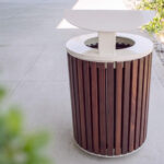 Outdoor trash can with vertical wooden slats and a white metal lid, placed on a sidewalk next to greenery and decorative rocks.