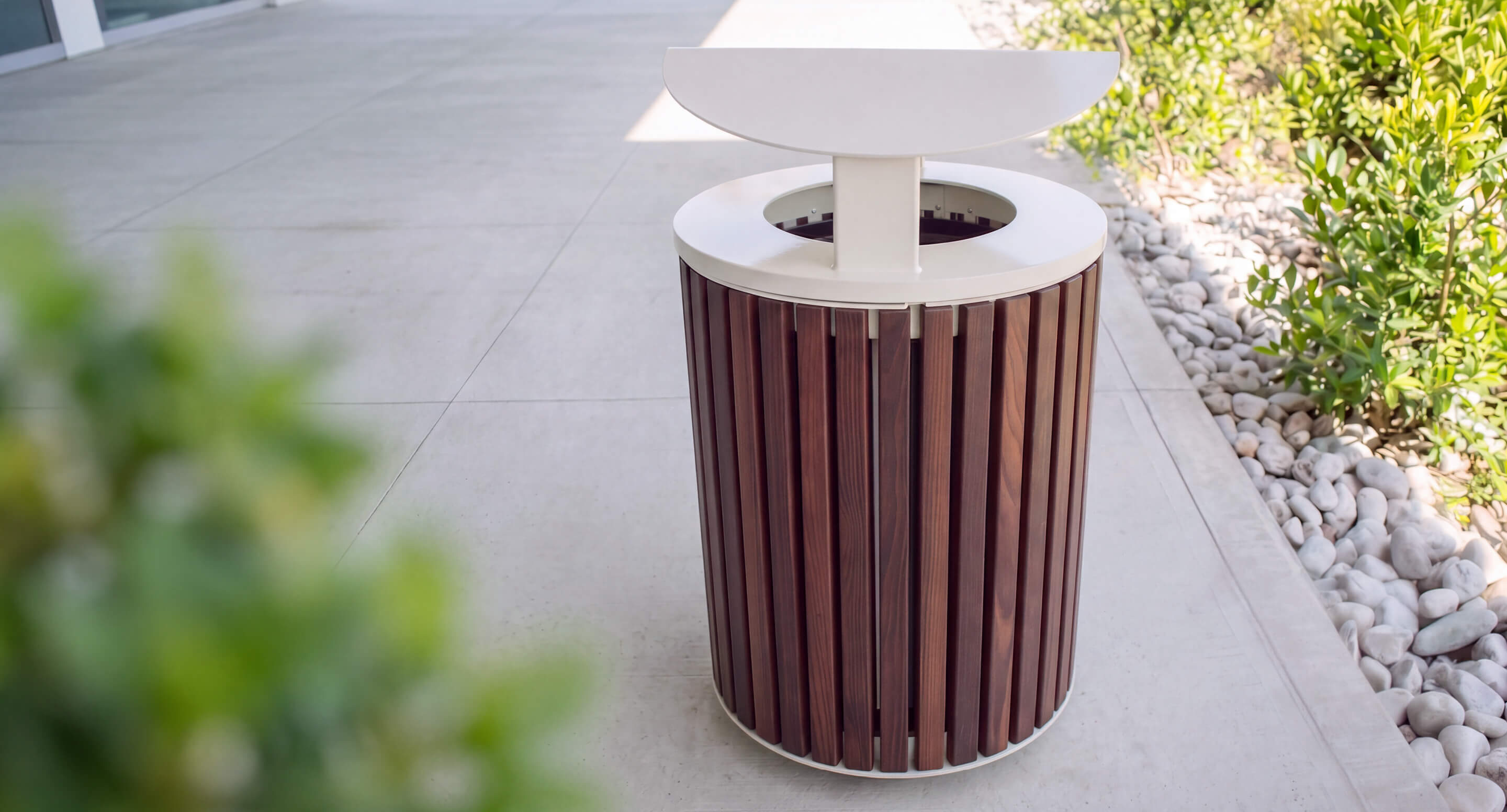 maglin_mtr-0720-trash-container_carousel-1 Outdoor trash can with vertical wooden slats and a white metal lid, placed on a sidewalk next to greenery and decorative rocks.